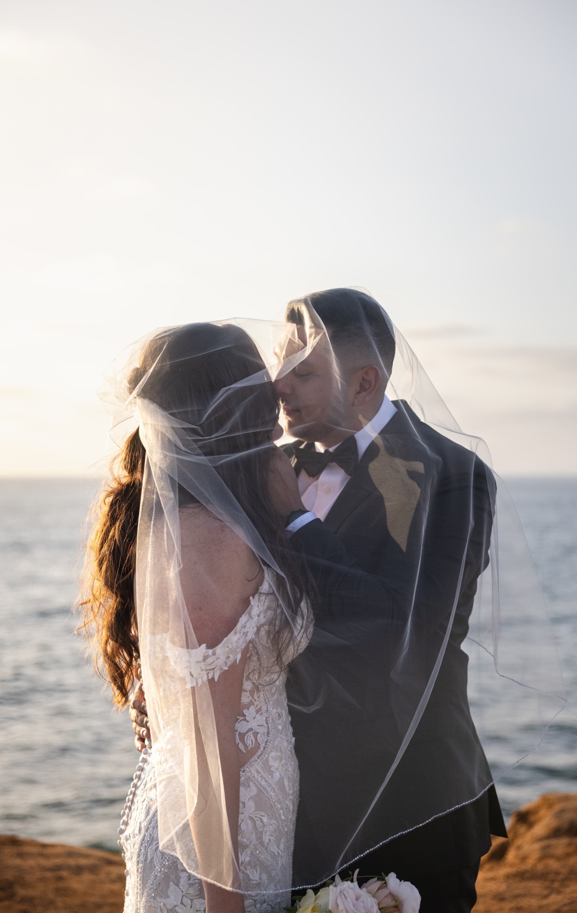 Bride and groom kissing under a flowing veil at sunset by the ocean