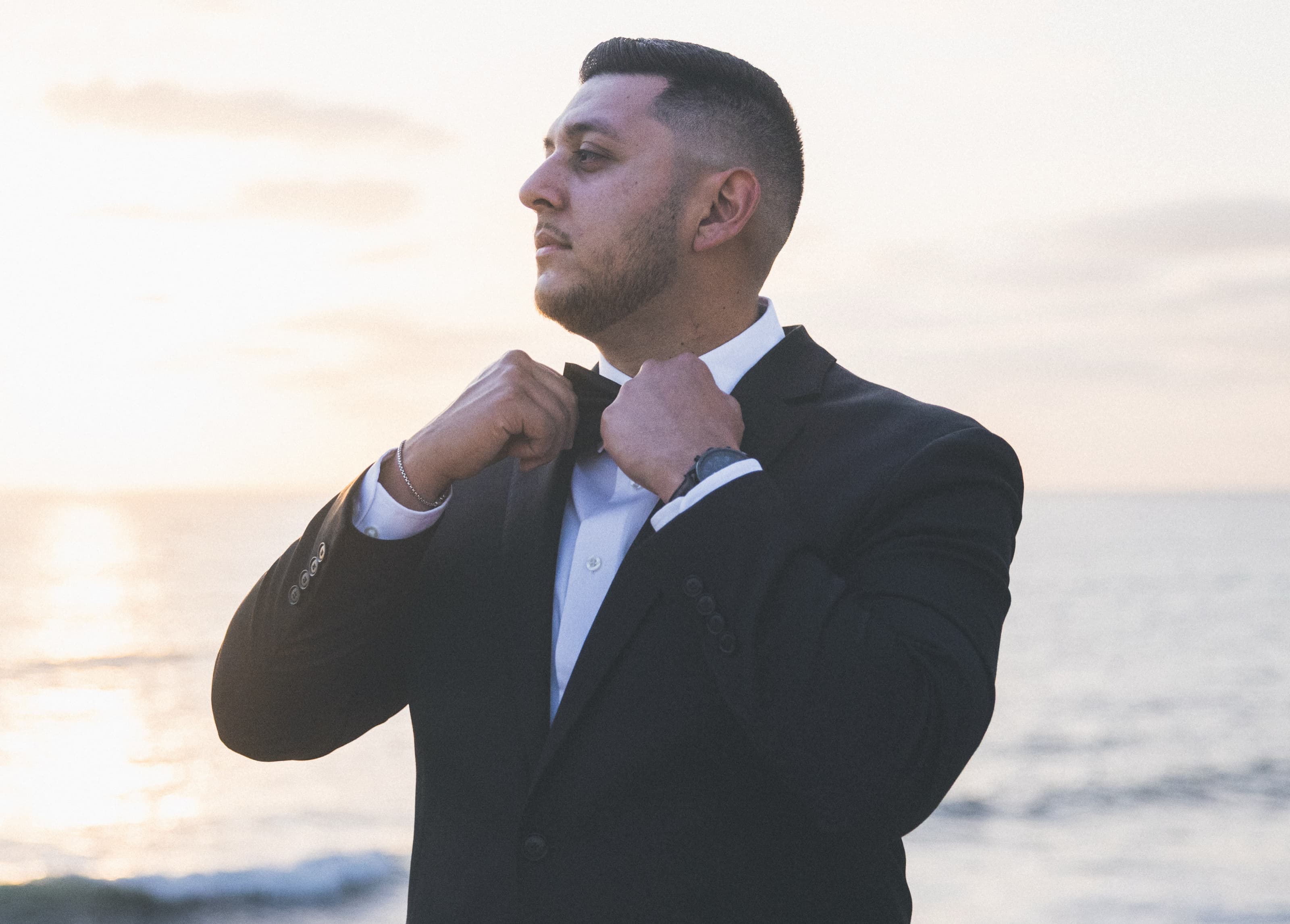 Groom adjusting his bow tie at sunset over the ocean