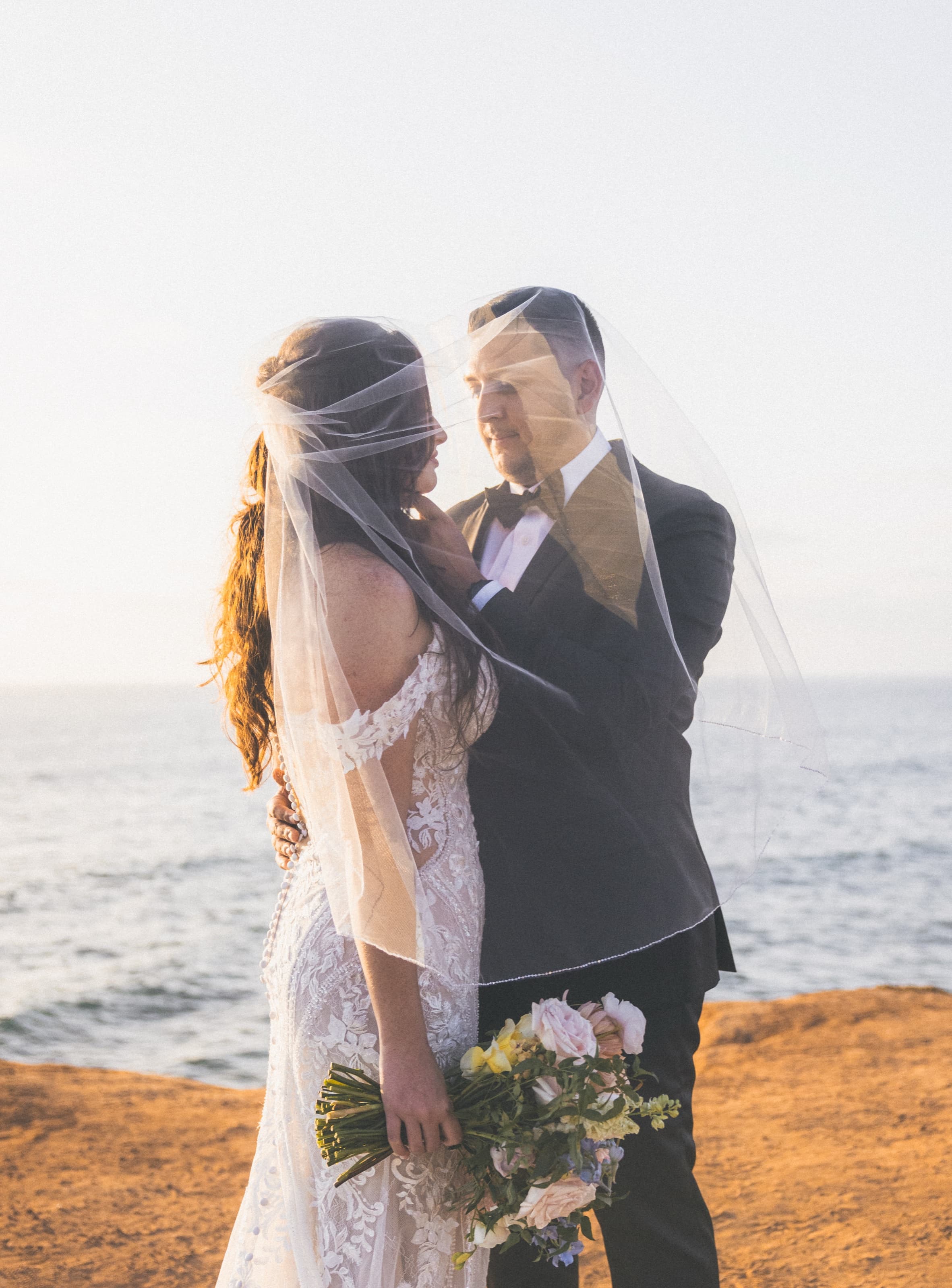 Bride with bouquet under a flowing veil with groom at sunset